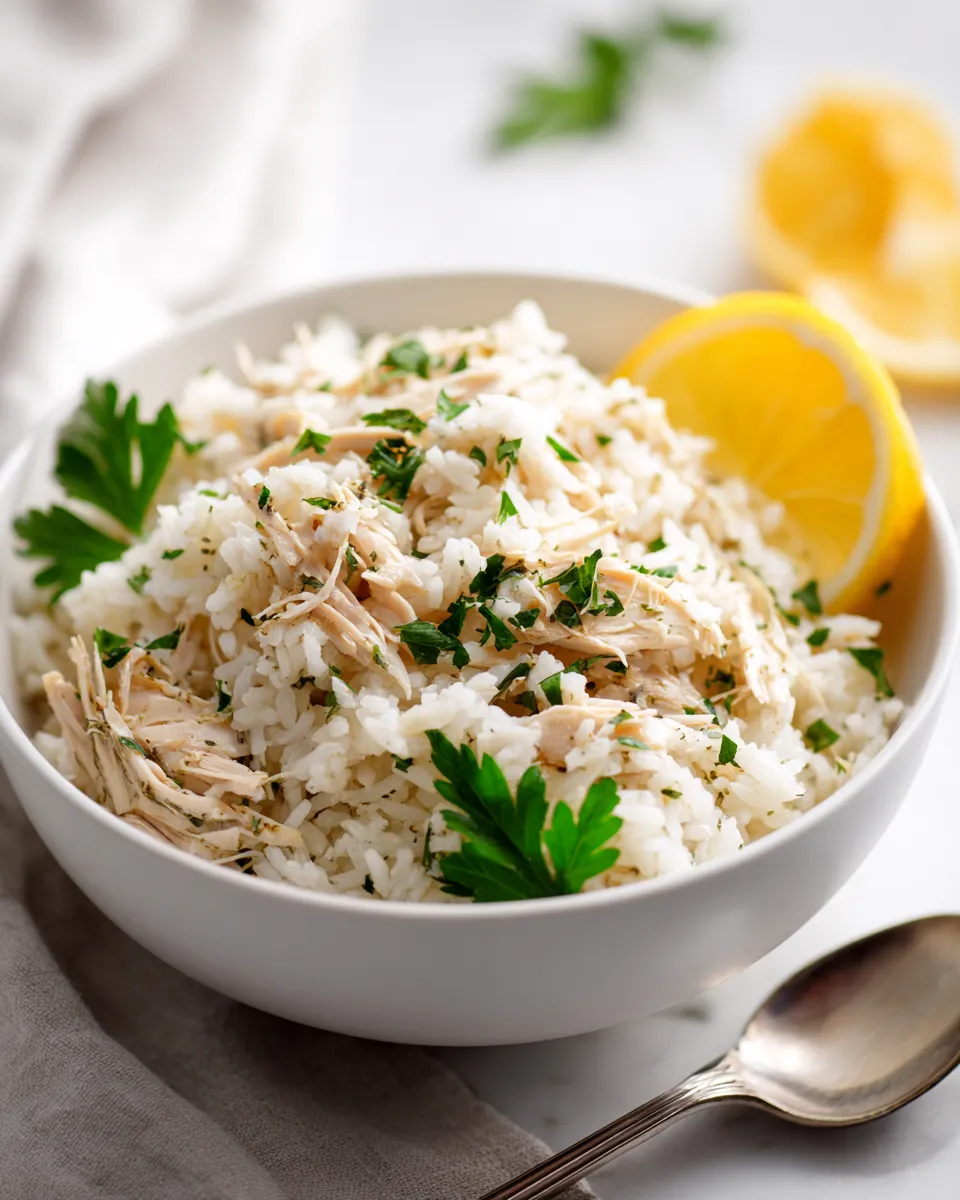 A close-up, realistic photo of slow cooker lemon herb chicken and rice served in a white bowl, showing fluffy rice and tender shredded chicken, garnished with fresh parsley and a lemon wedge.